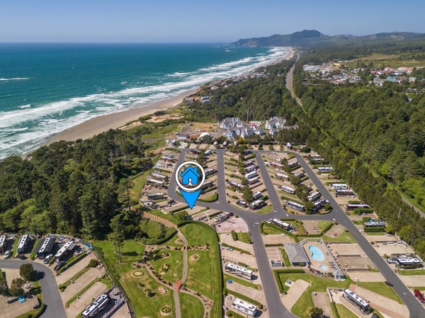 Aerial view of a coastal vacation rental resort nestled between forested hills and pristine oceanfront. The property features organized parking areas, pool facilities, and immediate access to the sandy beach.