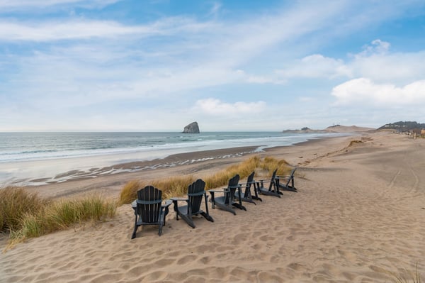 Expansive sandy beach with Adirondack chairs facing the ocean and dramatic rock formations in the distance.
