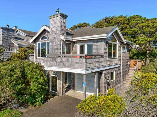 Charming coastal property featuring weathered shingle siding, a distinctive chimney tower, and lush green landscaping nestled among mature trees in a quiet neighborhood setting.