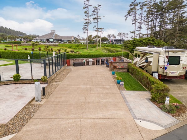 Spacious RV site with level parking, BBQ facilities, and lush landscaping surrounded by towering coastal pines and rolling green grounds.