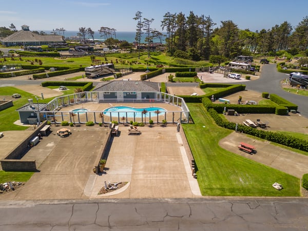 Aerial view reveals a resort property featuring an enclosed pool area surrounded by manicured lawns, mature pines, and sea views beyond. The landscaped grounds include parking areas and recreational spaces within a coastal setting.