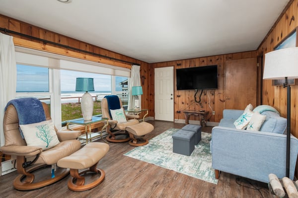 Relax in your cozy living room with warm wood-paneled walls, plush seating, and stunning ocean views just steps away—your perfect beachside escape awaits.