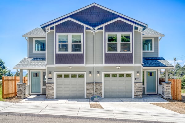 Modern duplex featuring charming coastal architecture with blue and gray tones, dual garages, and welcoming front entrances framed by stone accents.