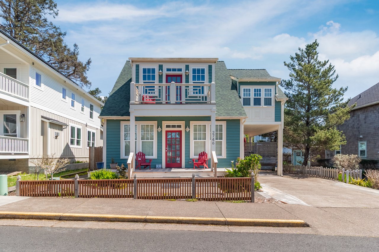 Charming coastal cottage with teal siding, red door, and welcoming front porch in peaceful neighborhood setting.