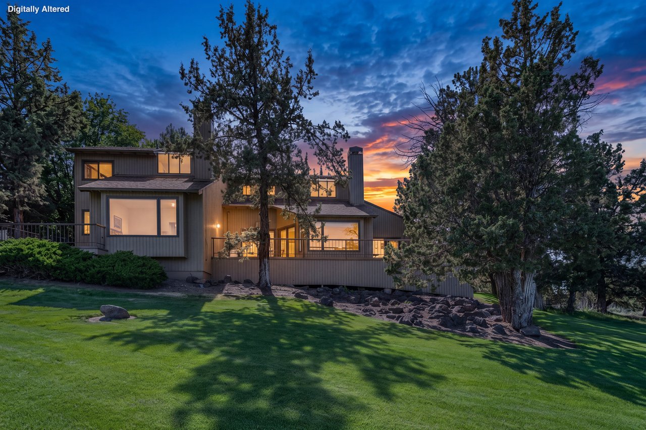 Modern retreat set on manicured lawns beneath towering pines, framed by vibrant sunset skies. The property offers a peaceful natural setting with mature trees and open green spaces.