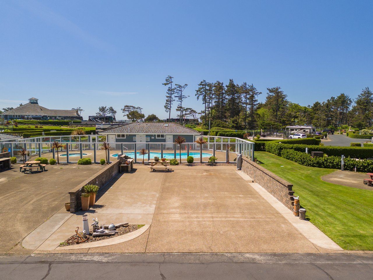 A spacious property featuring a pool, manicured lawns, picnic areas, and mature pine trees, set beneath clear blue skies in a tranquil coastal setting.