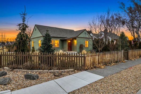 Charming green cottage with warm windows glowing against a stunning sunset sky, nestled in a quiet residential neighborhood.